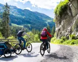 Zwei Radfahrer mit Rucksäcken und Anhänger fahren auf einer Bergstraße.