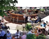 Gäste sitzen an Tischen und an der Theke eines Biergartens unter Sonnenschirmen.
