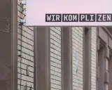 Leuchtreklame mit dem Schriftzug "WIR KOMPLIZEN" über einer Fassade.
