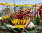 Eine rote Insektenfigur mit vielen Armen steht vor einer gelben Achterbahn.
